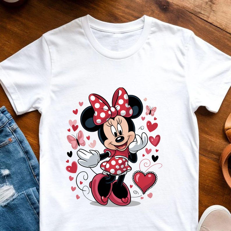 May include: A white short-sleeved t-shirt featuring a graphic print of Minnie Mouse wearing a red and white polka-dot dress and matching bow. The design is surrounded by various red and pink hearts and small butterflies on the front center.