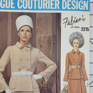 May include: A vintage sewing pattern for a double-breasted jacket and skirt suit. The pattern is from Vogue Couturier Design, Fabiani of Italy, and is number 2215. The model is wearing a white hat and a plaid jacket and skirt.