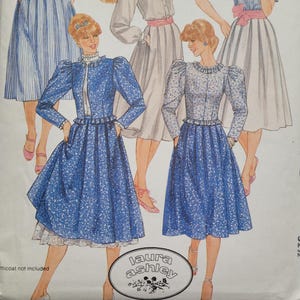 May include: A vintage Laura Ashley sewing pattern featuring a blue and white floral print dress with puffed sleeves and a gathered skirt. The pattern includes a separate white lace petticoat. The pattern is for sizes 8-16.