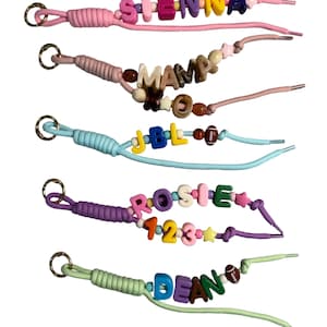May include: A collection of personalized keychains with colorful letter beads spelling out names and words. Each keychain features a different color scheme and includes a metal ring for attachment. The keychains are made with shoelaces.