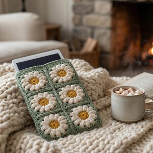 May include: A cozy scene featuring a tablet in a green and white daisy crochet case, resting on a cream-colored knitted blanket. A mug of hot chocolate and an open book sit nearby, with a fireplace in the background.