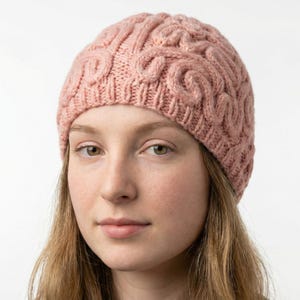 May include: A knitted beanie in a soft rose color. The hat features a cable knit design across the crown. The beanie is made of a soft, warm material, perfect for cold weather. The hat is shown on a person.