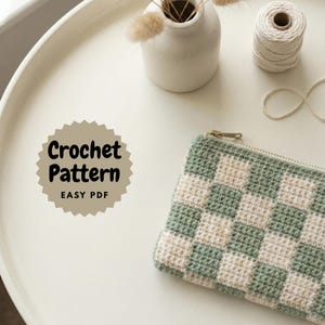 May include: A crochet pattern for a zippered pouch with a checkerboard design in light green and cream. The text "Crochet Pattern EASY PDF" is displayed on a circular label. A small white vase with dried flowers and a spool of white thread are also visible.