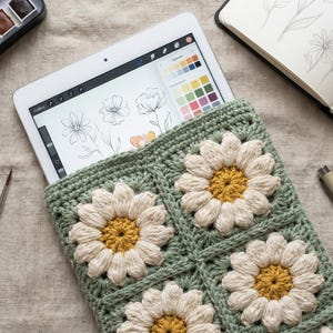 Puede incluir: Una funda de ganchillo verde salvia con cuatro motivos cuadrados con diseños de margaritas blancas y amarillas. La funda contiene una tableta con bocetos florales, rodeada de tres pinceles, un set de acuarelas, un cuaderno y un bolígrafo.