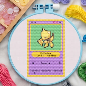 Könnte beinhalten: Kreuzstich-Stickerei einer Abra-Pokémon-Karte in einem blauen Stickrahmen. Die Karte zeigt einen gelben Abra-Charakter auf grünem Hintergrund mit Text wie "Psi Pokémon" und "Psyshock". Die Karte ist 89 cm lang und wiegt 19,5 kg.
