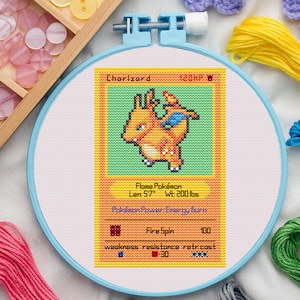 May include: A cross-stitch embroidery of a Charizard Pokémon card, set in a light blue hoop. The card features an orange Charizard with text that reads "Flame Pokémon", "Fire Spin", and "100". The card is 170 cm long and weighs 90 kg.