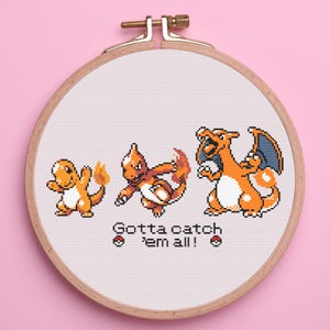 May include: Cross-stitch embroidery in a wooden hoop featuring three orange and white pixelated creatures. The text "Gotta catch 'em all!" is below the creatures. The background is white, and the hoop is set against a pink background.
