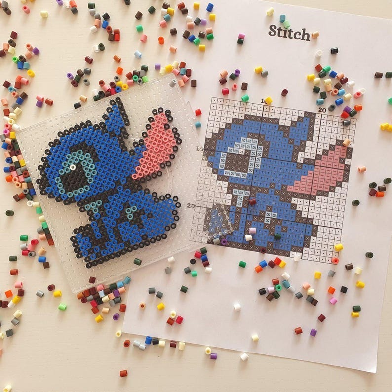 May include: A completed blue and pink bead art of the character Stitch, next to a pattern guide. The word "Stitch" is printed on the pattern guide. Scattered around are various colored beads, used for the art project.