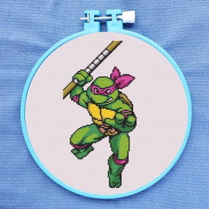 Op de afbeelding: Kruissteekpatroon van een Teenage Mutant Ninja Turtle die een katana-zwaard vasthoudt. De schildpad is groen met een paarse masker en bandana. De achtergrond is wit.
