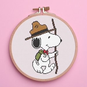 May include: A cross-stitch embroidery pattern of Snoopy dressed as a scout with a hat, backpack, and walking stick. The pattern is framed in a round wooden hoop.