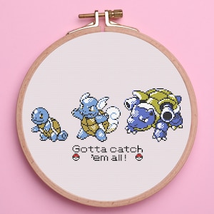 May include: Cross-stitch embroidery in a wooden hoop featuring three blue and green pixelated turtle-like creatures. The text "Gotta catch 'em all!" is below the creatures, with two red and white pokeball symbols.