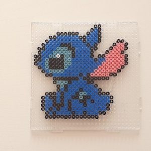 May include: A pixel art rendition of Stitch from Lilo & Stitch, crafted from blue, black, and pink beads. The artwork is set on a clear plastic pegboard, showcasing the character's distinctive features.