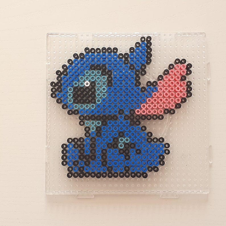 May include: A pixel art rendition of Stitch from Lilo & Stitch, crafted from blue, black, and pink beads. The artwork is set on a clear plastic pegboard, showcasing the character's distinctive features.
