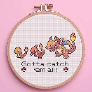 May include: Cross-stitch embroidery in a wooden hoop featuring three stages of a fire-type creature evolution. The text "Gotta catch 'em all!" is stitched below the creatures, with two Poké Ball designs.