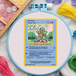 May include: Cross-stitch embroidery of a Squirtle Pokémon card. The design features a pixelated Squirtle character, text, and a blue border. The card reads "Squirtle" and has a length of 18 inches and a weight of 15 lbs.
