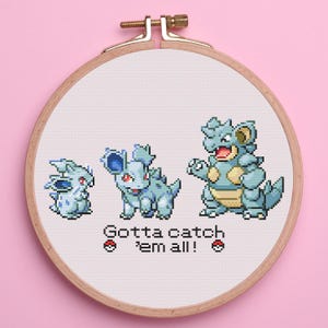 May include: A cross-stitch embroidery hoop featuring three blue and tan cartoon creatures. The text "Gotta catch 'em all!" is stitched below the creatures. The hoop is made of wood and is set against a pink background.