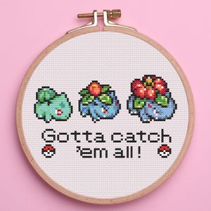 May include: Cross-stitch embroidery in a wooden hoop featuring three stages of a Bulbasaur evolution. The text "Gotta catch 'em all!" is stitched below the characters, with a Pokeball on each side. The background is light pink.