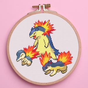 May include: A cross-stitch embroidery hoop featuring three colorful, pixelated fire-type creatures. The creatures are yellow, blue, and red, with flames emanating from their bodies. The hoop is made of wood and set against a pink background.