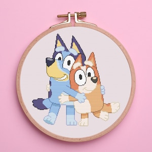 May include: A cross-stitch embroidery hoop featuring two cartoon dogs hugging. One dog is blue with a yellow muzzle, and the other is orange and white. The hoop is made of wood and has a brass clasp. The background is pink.
