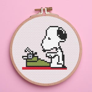 Puede incluir: Bordado de punto de cruz de un personaje de dibujos animados, Snoopy, sentado sobre una superficie verde con una máquina de escribir. El diseño está enmarcado en un aro de madera sobre un fondo rosa. El personaje es blanco con detalles negros.