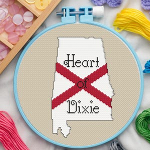May include: A cross-stitch embroidery hoop featuring the outline of the state of Alabama. The design includes a red cross and the words "Heart of Dixie" in black. The hoop is light blue, and the fabric is beige.