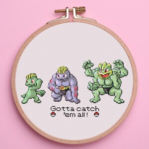 May include: Cross-stitch embroidery in a wooden hoop featuring three pixelated Pokémon characters in green and purple. The text "Gotta catch 'em all!" is below the characters. The background is pink.