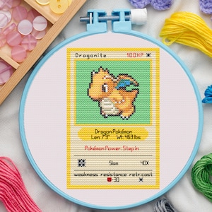 May include: A cross-stitch embroidery hoop featuring a Dragonite Pokémon card design. The card includes the name "Dragonite", HP, and other game details. The design is in a light blue hoop, with colourful threads and craft supplies in the background.