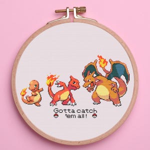 May include: A cross-stitch embroidery hoop featuring the evolution of a fire-type Pokémon. The design shows three stages of the creature in shades of orange, red, and yellow, with the text "Gotta catch 'em all!" in black.