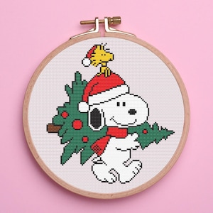 Puede incluir: Snoopy, el querido beagle de Peanuts, lleva un gorro de Papá Noel rojo y sostiene un árbol de Navidad verde. El mejor amigo de Snoopy, Woodstock, está posado en la parte superior de su gorro.