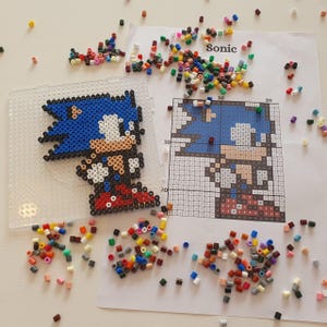 Può includere: Una creazione di pixel art di Sonic the Hedgehog, realizzata con perline di plastica colorate. Accanto, una base forata e uno schema stampato. Perline sparse di vari colori circondano l'opera, pronte per l'uso. La parola "Sonic" è stampata sullo schema.
