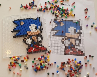 May include: A completed Sonic the Hedgehog pixel art creation made with colorful plastic beads, next to a pegboard and a printed pattern guide. Scattered beads in various colors surround the artwork, ready for use. The word "Sonic" is printed on the pattern.