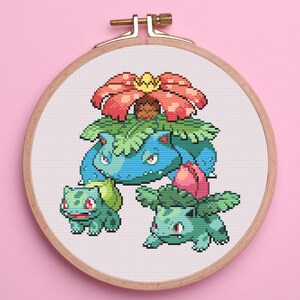 May include: Cross-stitch embroidery of a Bulbasaur family, featuring a large blue Venusaur with a red and yellow flower, and two smaller Bulbasaurs. The design is set in a wooden embroidery hoop against a pink background.