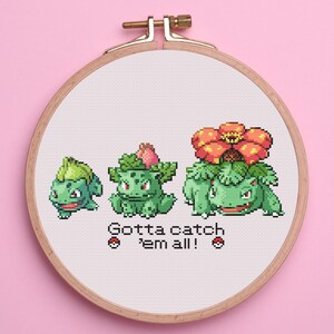 May include: A cross-stitch embroidery hoop featuring three green Pokémon characters in various stages of evolution. The text "Gotta catch 'em all!" is stitched below the characters, with a pink background.