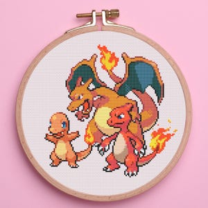 May include: A cross-stitch embroidery featuring three Pokémon characters: Charmander, Charmeleon, and Charizard. The design is set in a wooden embroidery hoop against a pink background. The characters are rendered in orange, red, and yellow.
