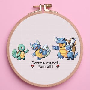 May include: Cross-stitch embroidery in a wooden hoop featuring three blue and yellow Pokémon characters. The text "Gotta catch 'em all!" is stitched below the characters, with two Poké Ball symbols.