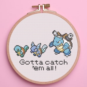 May include: A cross-stitch embroidery hoop featuring three blue and tan Pokémon characters in various stages of evolution. The text "Gotta catch 'em all!" is stitched below the characters. The hoop is made of wood and is set against a pink background.