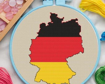 May include: A cross-stitch embroidery hoop depicting the map of Germany, coloured in the national flag's black, red, and yellow. The embroidery is set in a light blue hoop, with craft supplies such as thread and buttons in the background.