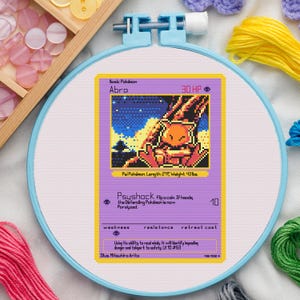 Könnte beinhalten: Kreuzstich-Stickerei einer Pokémon-Karte mit Abra. Die Karte ist lila mit einem pixeligen Bild von Abra. Der Text auf der Karte enthält "Abra", "Psyshock" und andere Spieldetails. Die Stickerei befindet sich in einem blauen Stickrahmen.