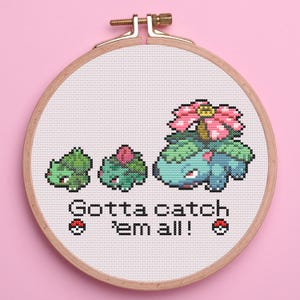 May include: Cross-stitch embroidery in a wooden hoop featuring the evolution of a Bulbasaur. The design includes the text "Gotta catch 'em all!" with two pixelated Pokeball icons. The background is a light pink.