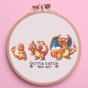 Può includere: Ricamo a punto croce in un telaio di legno con tre personaggi di cartoni animati arancioni e rossi con fiamme. Il testo "Gotta catch 'em all!" è sotto i personaggi. Lo sfondo è bianco e il telaio è posto su uno sfondo rosa.