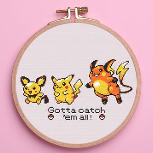 May include: Cross-stitch embroidery of three Pokémon characters in yellow and orange, framed in a wooden hoop. The text "Gotta catch 'em all!" is stitched below the characters, with two Poké Ball symbols. The background is light pink.