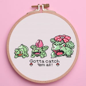 May include: Cross-stitch embroidery in a wooden hoop featuring three stages of a green Pokémon character evolution. The text "Gotta catch 'em all!" is stitched below the characters. The background is a light pink.