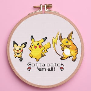 May include: Cross-stitch embroidery featuring three Pokémon characters: Pichu, Pikachu, and Raichu, with a lightning bolt. The text "Gotta catch 'em all!" is stitched below the characters. The embroidery is set in a wooden hoop.