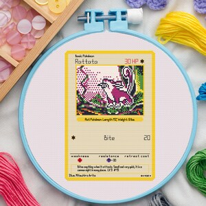 May include: A cross-stitch of a Rattata Pokémon card in a blue embroidery hoop. The card features a purple and white creature with the text "Rattata" and "Bite". The card is 25.4 cm long and weighs 3.6 kg.