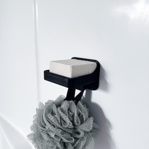 May include: A black soap dish with a bar of soap and a gray loofah hanging below. The soap dish is mounted on a white wall. The loofah is a large, round, mesh sponge.