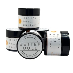 May include: Four small jars of Hill's Bull Butter, a skin care product. The jars are stacked on top of each other. The jars are black with white labels. The labels have the words "Hill's Bull Butter" and "Better Bull" on them. The labels also have a small orange slice graphic on them.