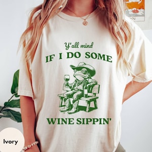 A woman wearing an ivory-colored T-shirt featuring a trendy funny slogan, Y'all Mind If I Do Some Wine Sippin', along with a retro graphic of a cowboy frog sitting on a bench and holding a glass of wine in his hand. The design is in darker green.
