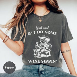 May include: Pepper-colored t-shirt with a white graphic of a frog in a cowboy hat, boots, and a wine glass, seated on a bench. Text reads "Y'all mind IF I DO SOME WINE SIPPIN'."