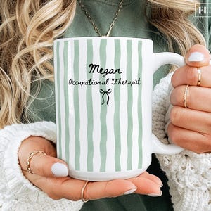 May include: A white ceramic mug featuring vertical sage green stripes. The text "Megan" appears above "Occupational Therapist" in black script and print, followed by a small black bow icon. The mug has a curved handle and a smooth finish.