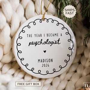 May include: A round white ceramic ornament featuring a black scalloped border and the text THE YEAR I BECAME A psychologist, followed by a small heart, the name MADISON, and the year 2026. The ornament hangs from a thin gold string.